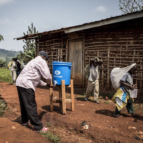 A man washes his hands under a bucket with a tap outside as people walk past.