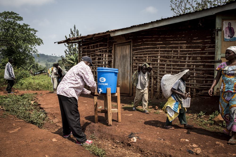 A man washes his hands under a bucket with a tap outside as people walk past.