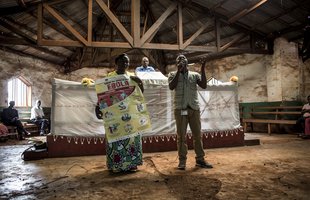Two public health promotors stand in a town hall holding a poster about Ebola and talking to an audience of community members about stopping the spread
