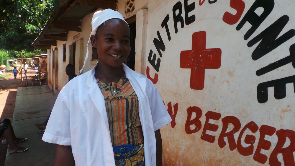 Solange wears a white nurse's uniform and stands next to a building with a red cross on it