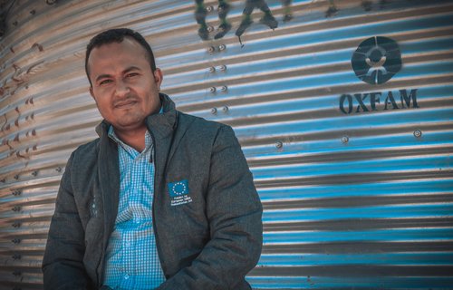 A portrait of Tamim next to an metal cylindrical Oxfam water tank