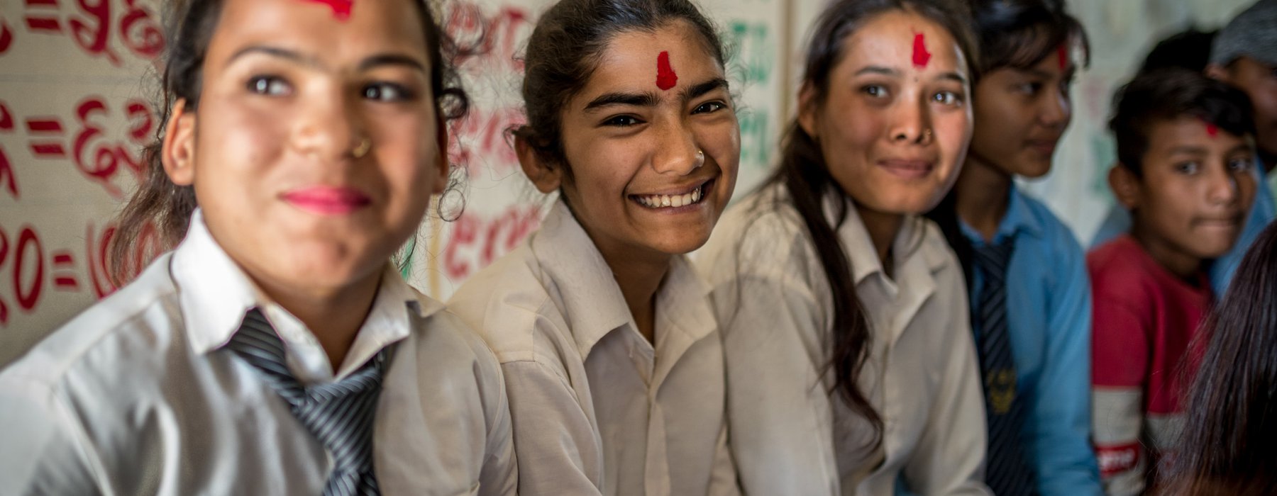 Three girls in a classroom setting smile at the camera. They have red powder on their foreheads.