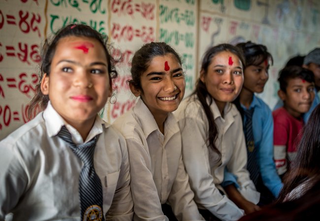 Three girls in a classroom setting smile at the camera. They have red powder on their foreheads.