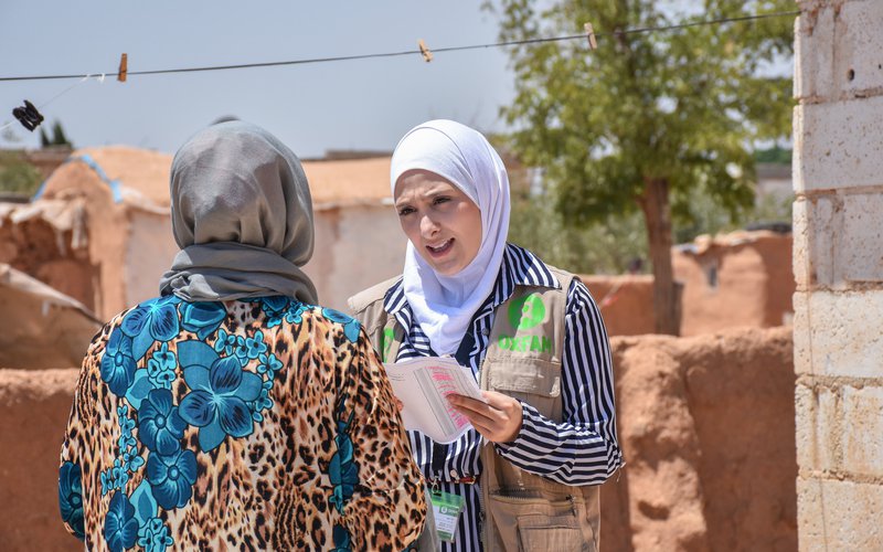 Sana wears a white hijab and Oxfam vest as she talks with a person in Rural Aleppo.