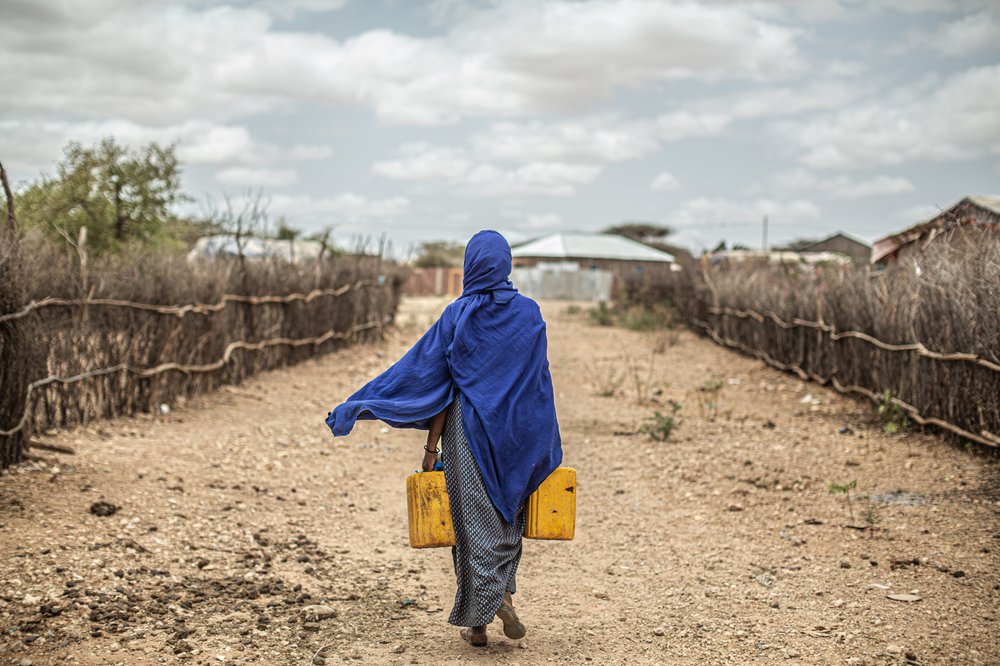 Amina wears cobalt blue and walks away down a dry path carrying two yellow water containers.