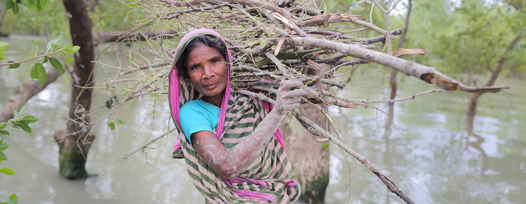 Nurjahan stands in floodwater with a bundle of sticks on her back and mud on her hands and smiles