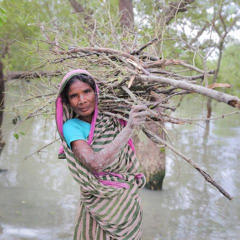 Nurjahan stands in floodwater with a bundle of sticks on her back and mud on her hands and smiles