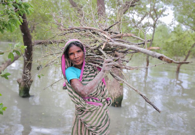 Nurjahan stands in floodwater with a bundle of sticks on her back and mud on her hands and smiles