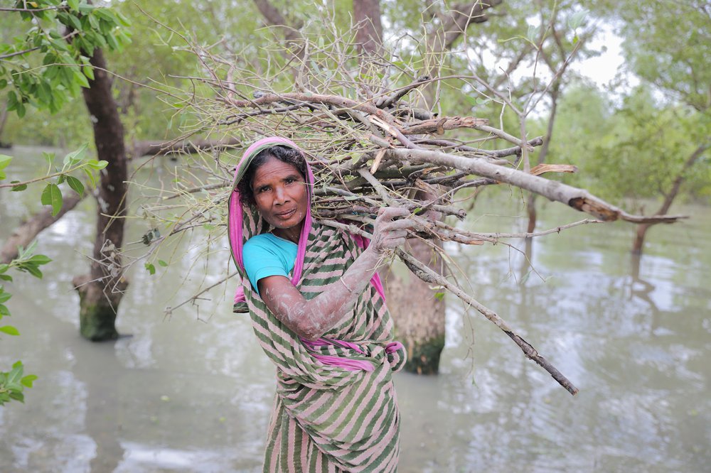 Nurjahan stands in floodwater with a bundle of sticks on her back and mud on her hands and smiles