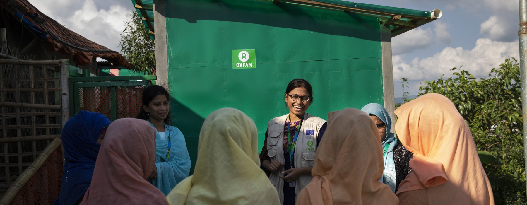 Oxfam staff member Iffat talks with a group of women wearing pastel-coloured headscarves about the toilets in the background