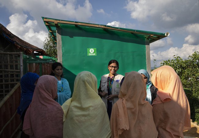 Oxfam staff member Iffat talks with a group of women wearing pastel-coloured headscarves about the toilets in the background