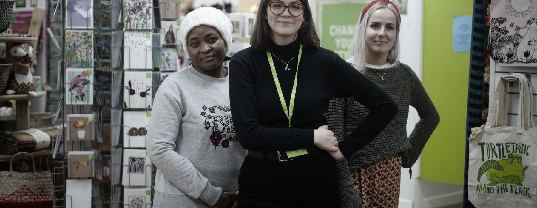 Three Oxfam shop volunteers wearing green lanyards stand outside an Oxfam shop