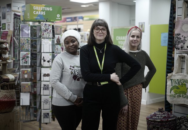 Three Oxfam shop volunteers wearing green lanyards stand outside an Oxfam shop