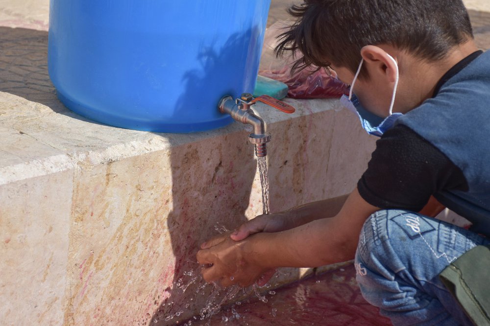 An eight-year-old boy wearing a face mask washes his hands under a tap.