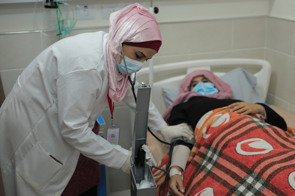 Nurse Heba Shalan is wearing a facemask and stethoscope and bending over a patient in a hospital bed who also wears a mask