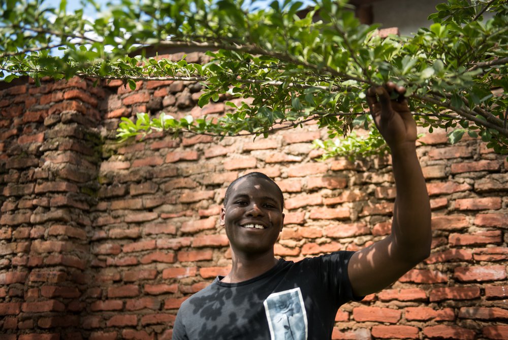 Isaac smiles and holds on to the branch of a tree by a red brick wall in Malawi
