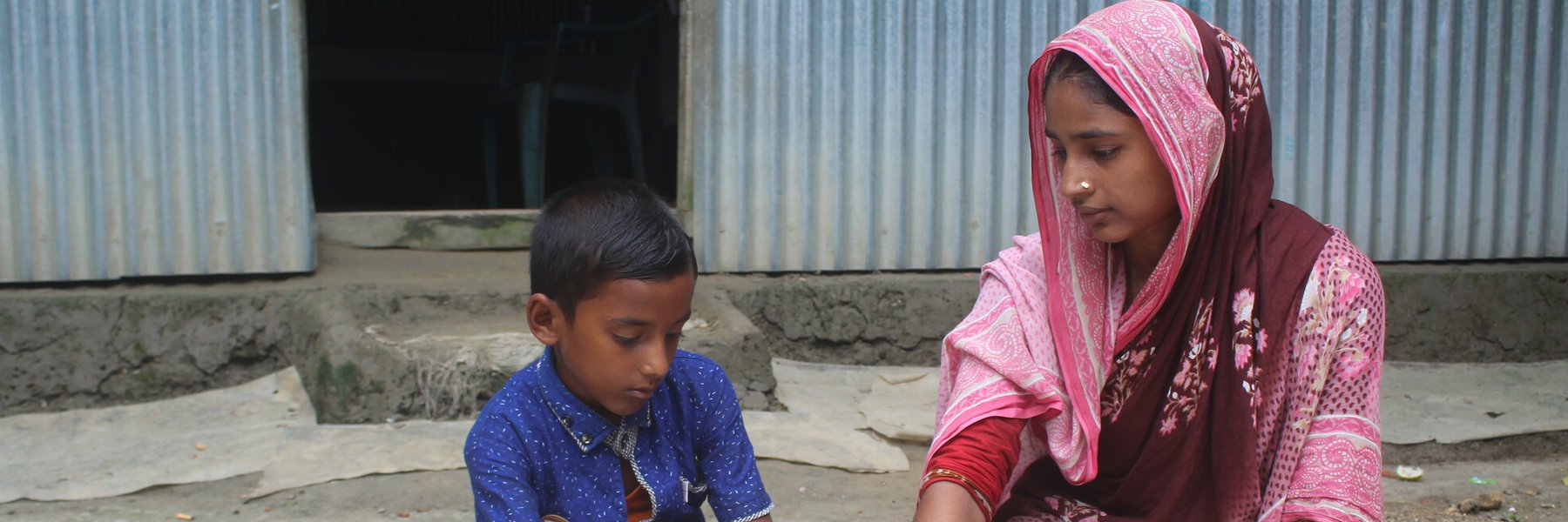 Shabana and her son sit in front of their home on the ground looking at a textbook together