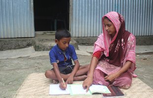 Shabana and her son sit in front of their home on the ground looking at a textbook together