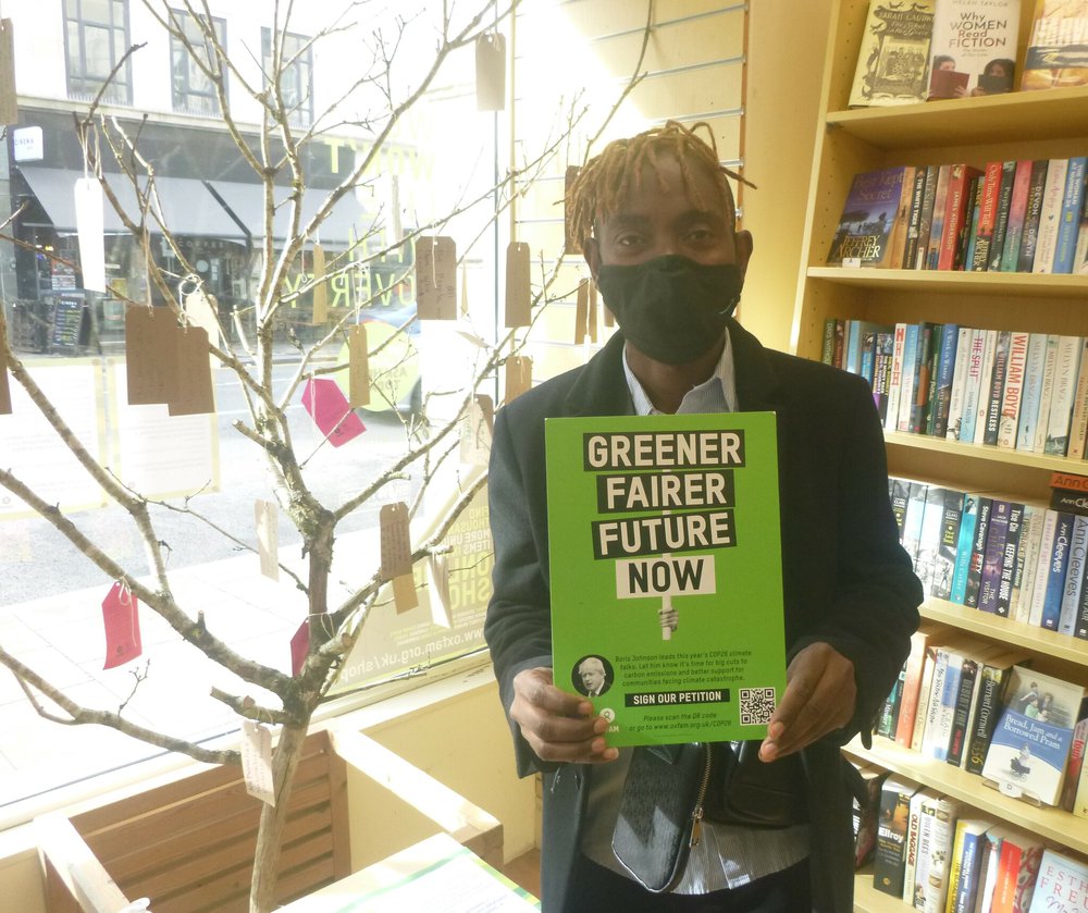 Oxfam shop volunteer Otis holds a printed sign that says 'greener, fairer future now' on it