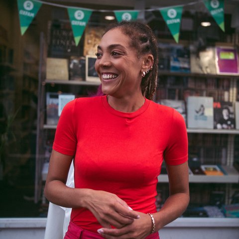 Miquita Oliver smiling outside an Oxfam shop wearing a second hand bright red T-shirt