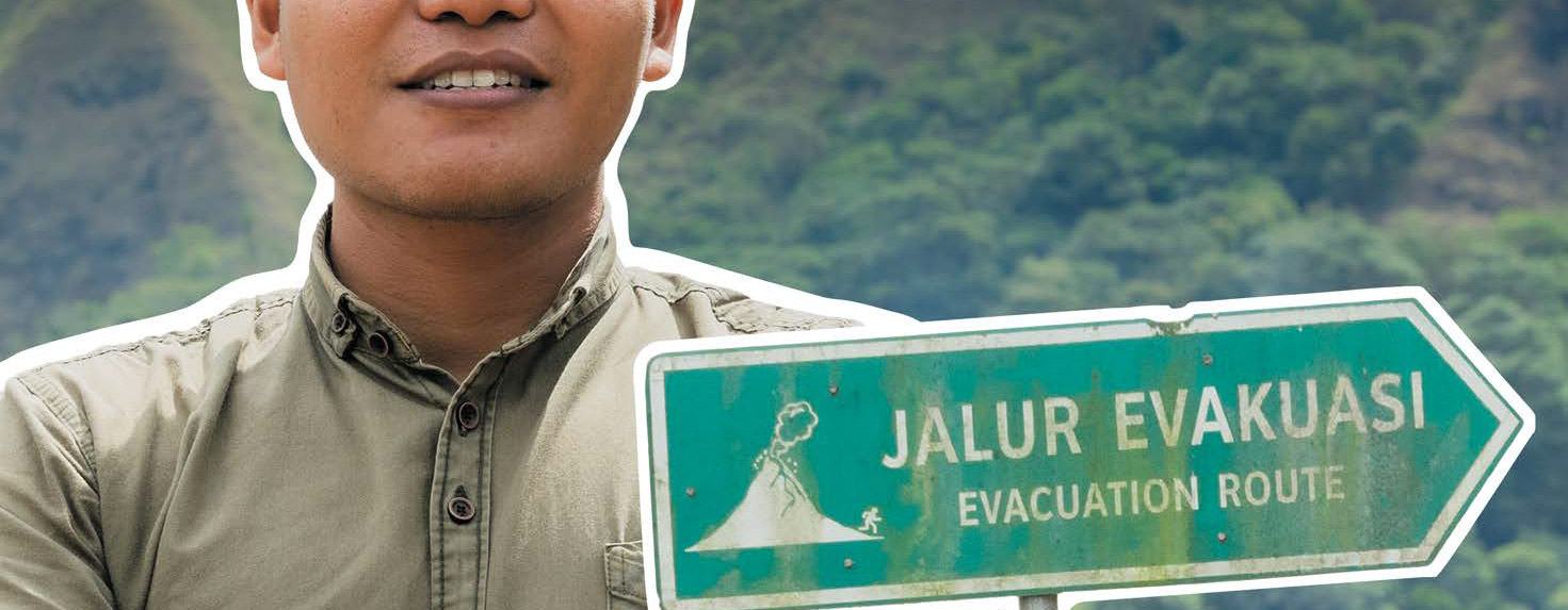 A smiling man with folded arms and a signpost reading 'Jalur Evakuasi Evacuation Route'