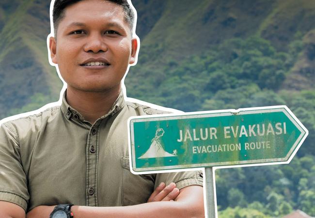 A smiling man with folded arms and a signpost reading 'Jalur Evakuasi Evacuation Route'