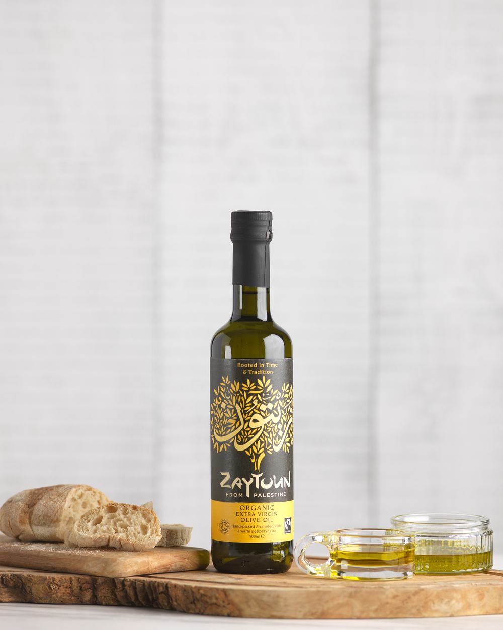 A bottle of Zaytoun olive oil on a bread board beside slices of bread and glass ramekins of oil.