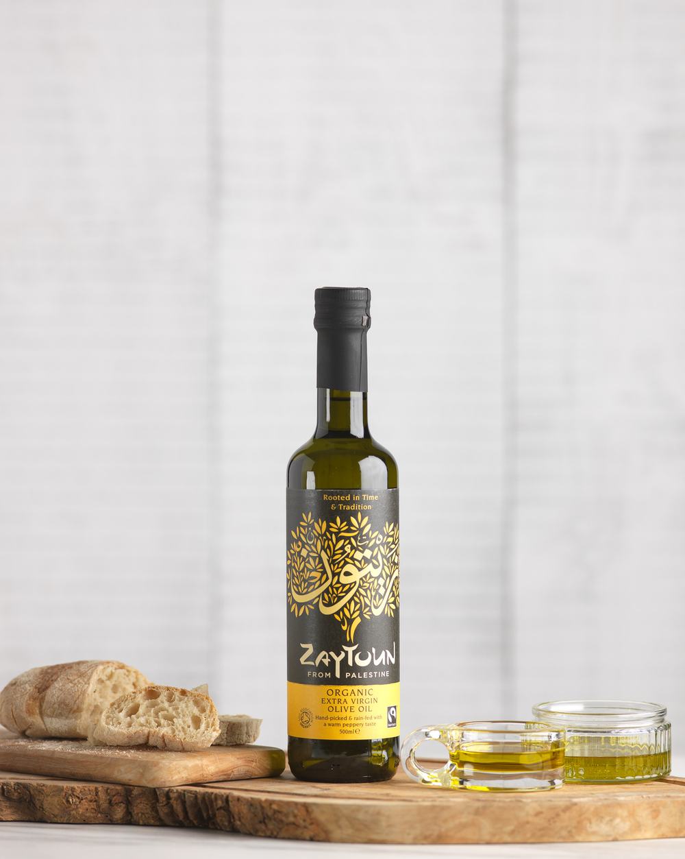 A bottle of Zaytoun Extra Virgin Olive Oil displayed on a wooden board alongside bread and dipping dishes of olive oil.