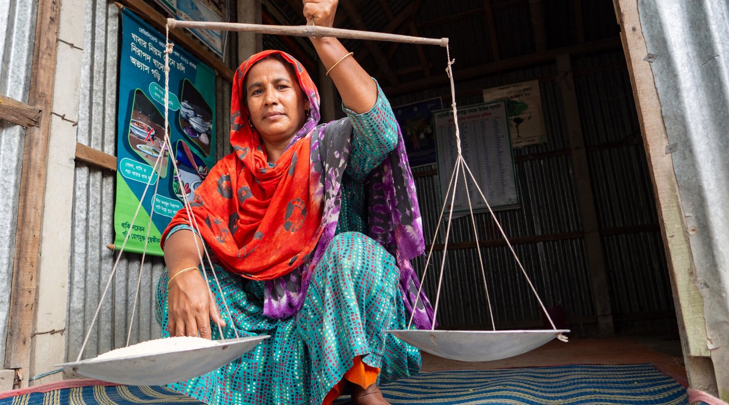 A woman holding up a pair of old-fashioned scales, balancing a bowl on either side. One of the two bowls is full of rice.