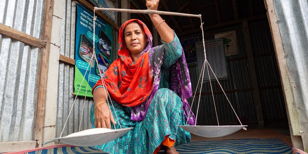 A woman holding up a pair of old-fashioned scales, balancing a bowl on either side. One of the two bowls is full of rice.