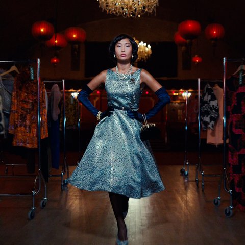 An Asian model in her early 20s wears a silver dress with a cinched waist and full skirt as well as long sleeved navy gloves and some glitzy accessories.