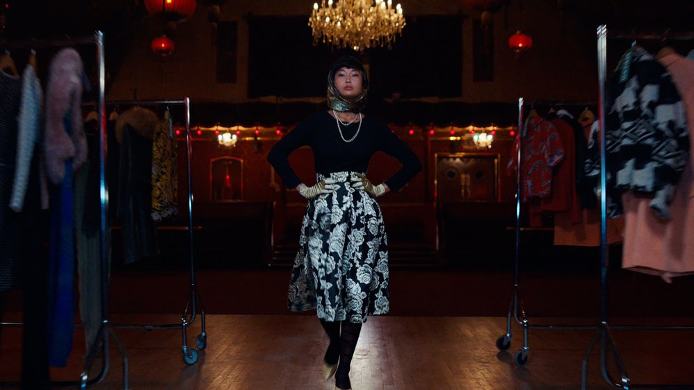 An Asian model in her early 20s wears a black and white floral skirt, black top and a silver scarf on her head. As well as a string of pearls.