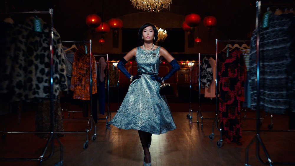 An Asian model in her early 20s wears a silver dress with a cinched waist and full skirt as well as long sleeved navy gloves and some glitzy accessories.