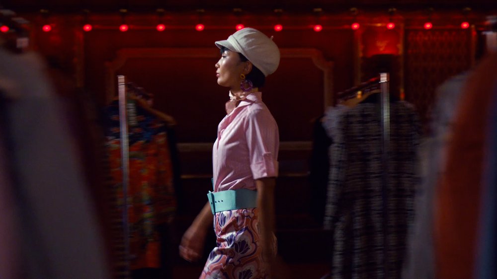 An Asian model in her early 20s wears a white cap and a pink shirt with a chunky pale blue belt and some pattered pink trousers and stands in profile.