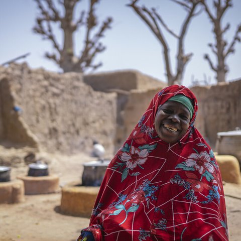 Mariam is smiling, wearing a red tunic and head covering with a pattern of white and blue flowers standing in front cooking firepits outside of sandy walls and buildings