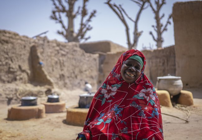 Mariam is smiling, wearing a red tunic and head covering with a pattern of white and blue flowers standing in front cooking firepits outside of sandy walls and buildings