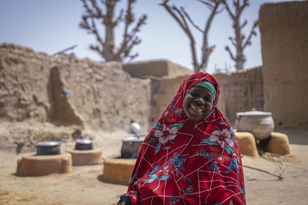 Mariam is smiling, wearing a red tunic and head covering with a pattern of white and blue flowers standing in front cooking firepits outside of sandy walls and buildings