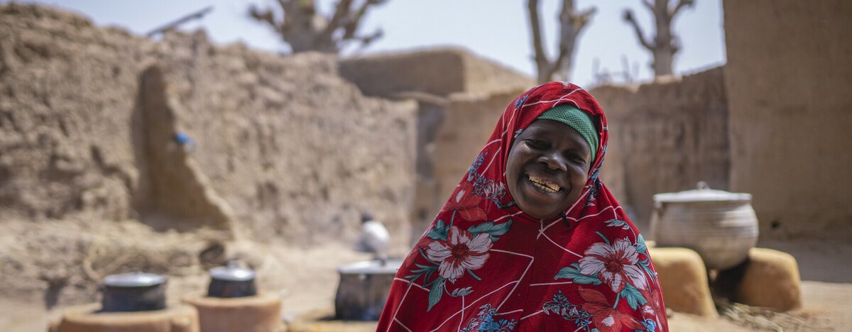 Mariam is smiling, wearing a red tunic and head covering with a pattern of white and blue flowers standing in front cooking firepits outside of sandy walls and buildings