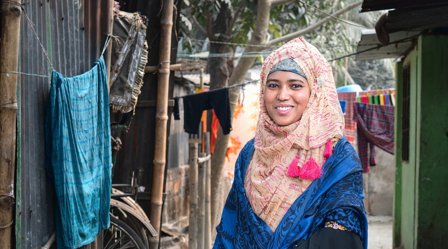 Rima, a young woman from Bangladesh smiles towards the camera while wearing a colourful dress and headscarf.
