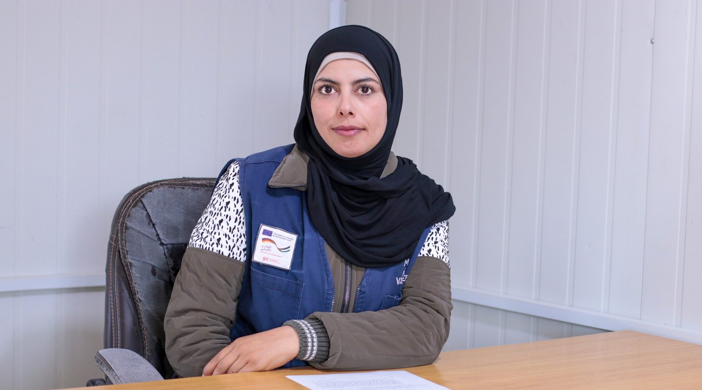 Hamda wears a black hijab and blue work vest and sits at a desk with a laptop closed on top of it.