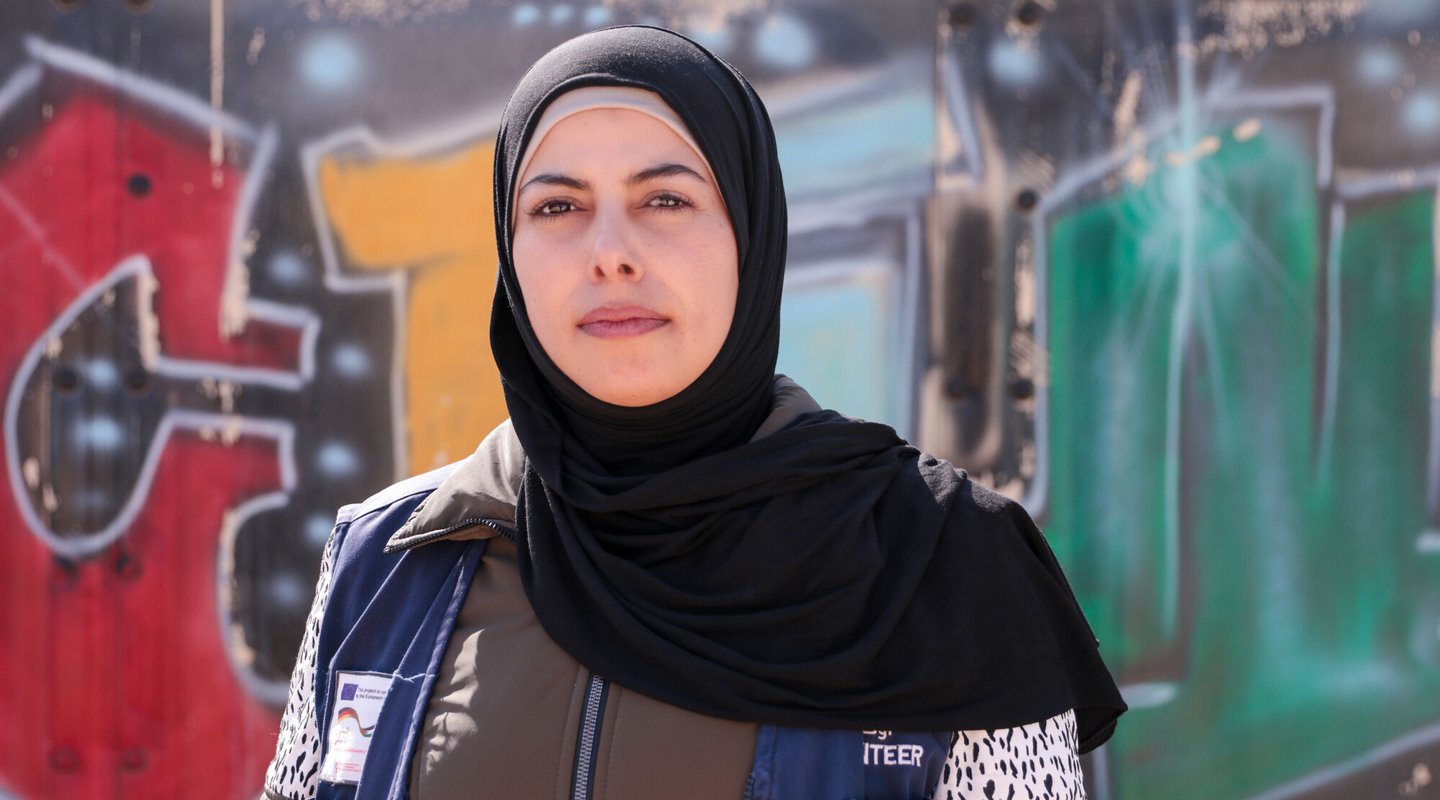 Hamda wears a black hijab and blue work vest and there is graffiti behind her