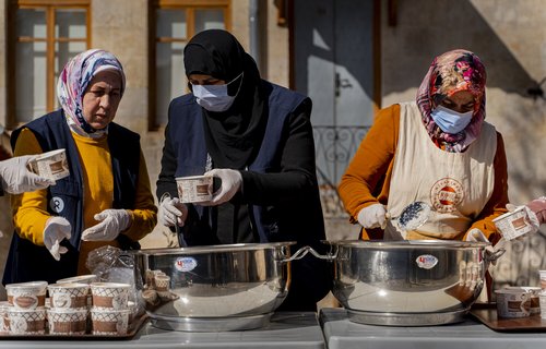 Three women in hijab wearing covid-safe masks with work vests on prepare food in big pans.