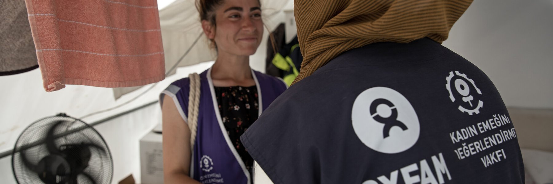 Two women wearing Oxfam KEDV vests inside a tent talking.