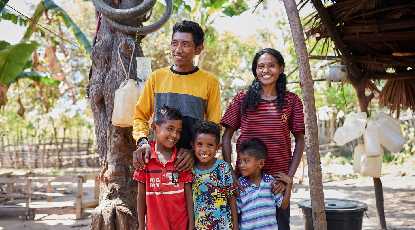 A smiling wife and husband stand with their three children outside their home in Timor-Leste.