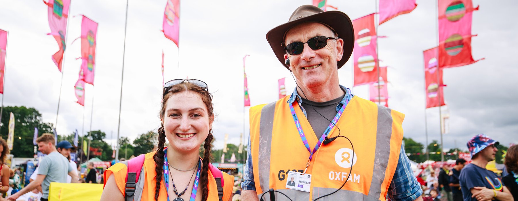 Oxfam Festival Stewards, Kat Atthey and Patrick O'Donnell smile at the camera while wearing Oxfam lanyards and orange visibility tabards at Womad Festival 2023.