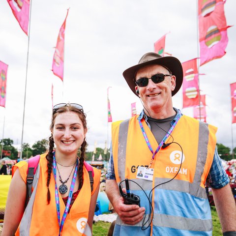 Oxfam Festival Stewards, Kat Atthey and Patrick O'Donnell smile at the camera while wearing Oxfam lanyards and orange visibility tabards at Womad Festival 2023.