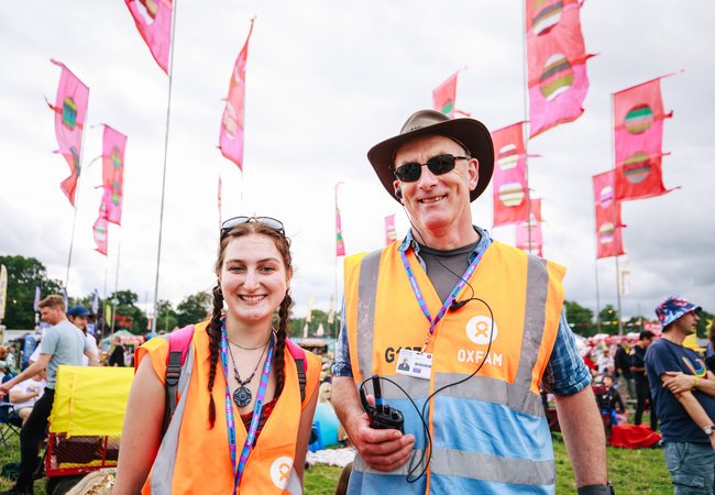 Oxfam Festival Stewards, Kat Atthey and Patrick O'Donnell smile at the camera while wearing Oxfam lanyards and orange visibility tabards at Womad Festival 2023.