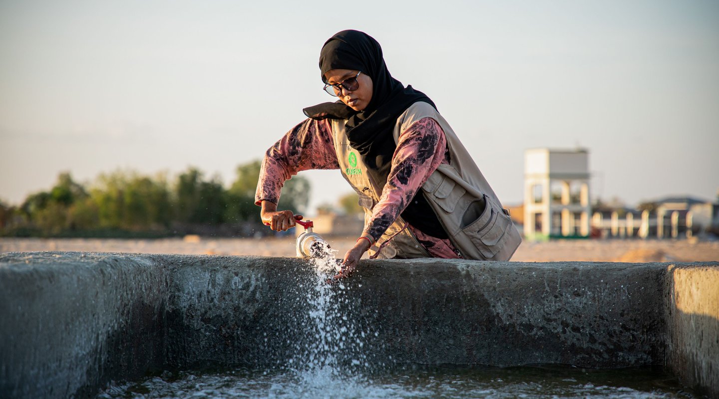 Person filling a plastic bottle with water from a concrete basin outdoors, with trees and a building visible in the background.