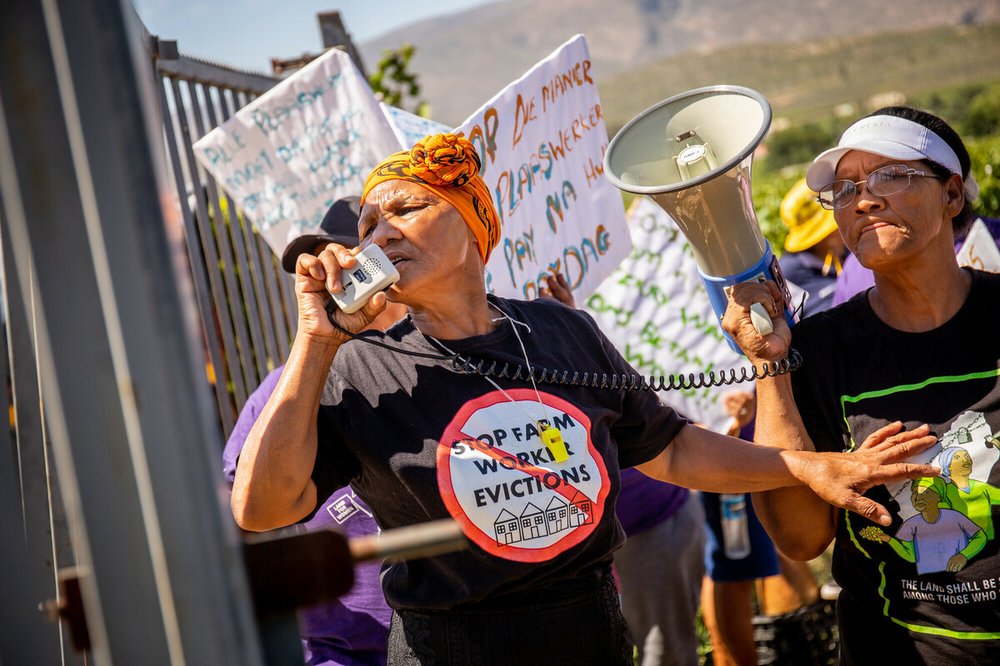 A group of protestors by iron railings. One protestor is speaking into a megaphone, and wearing a t-shirt that says "Stop farm worker evictions". Others are holding placards.