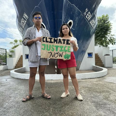 A man and a woman are standing in front of a monument that is the bow of a large boat. They are holding a handmade sign that says "Climate justice now".